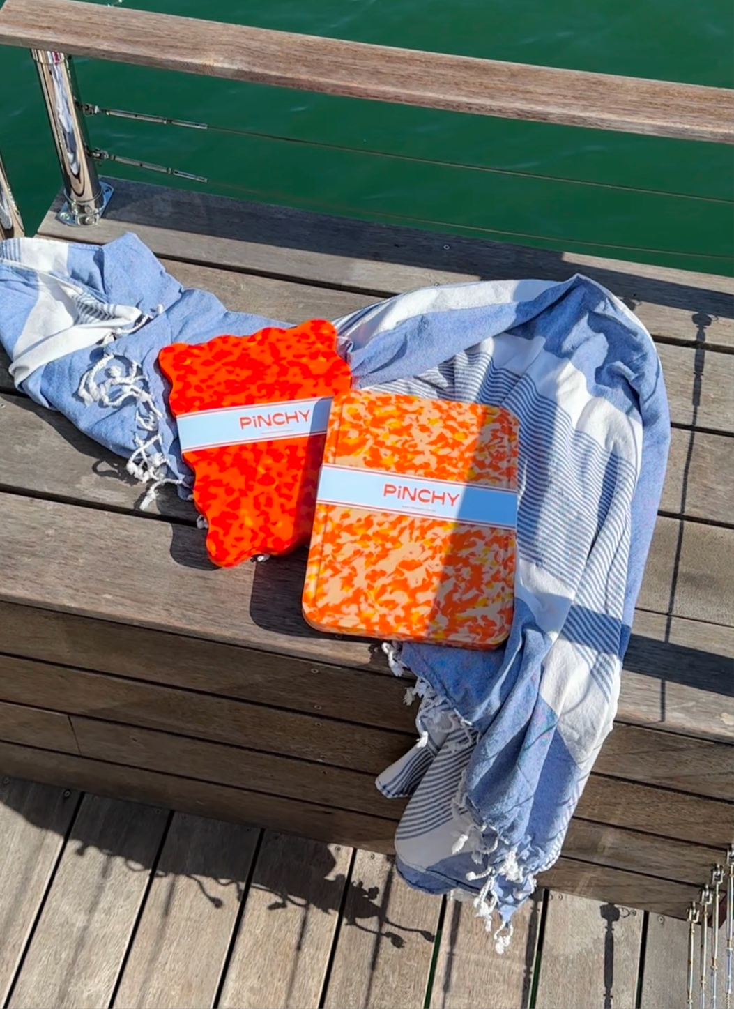 Two orange Pinchy chopping boards with wavy and rectangular shapes resting on a timber deck beside the ocean, styled with a blue striped towel.
