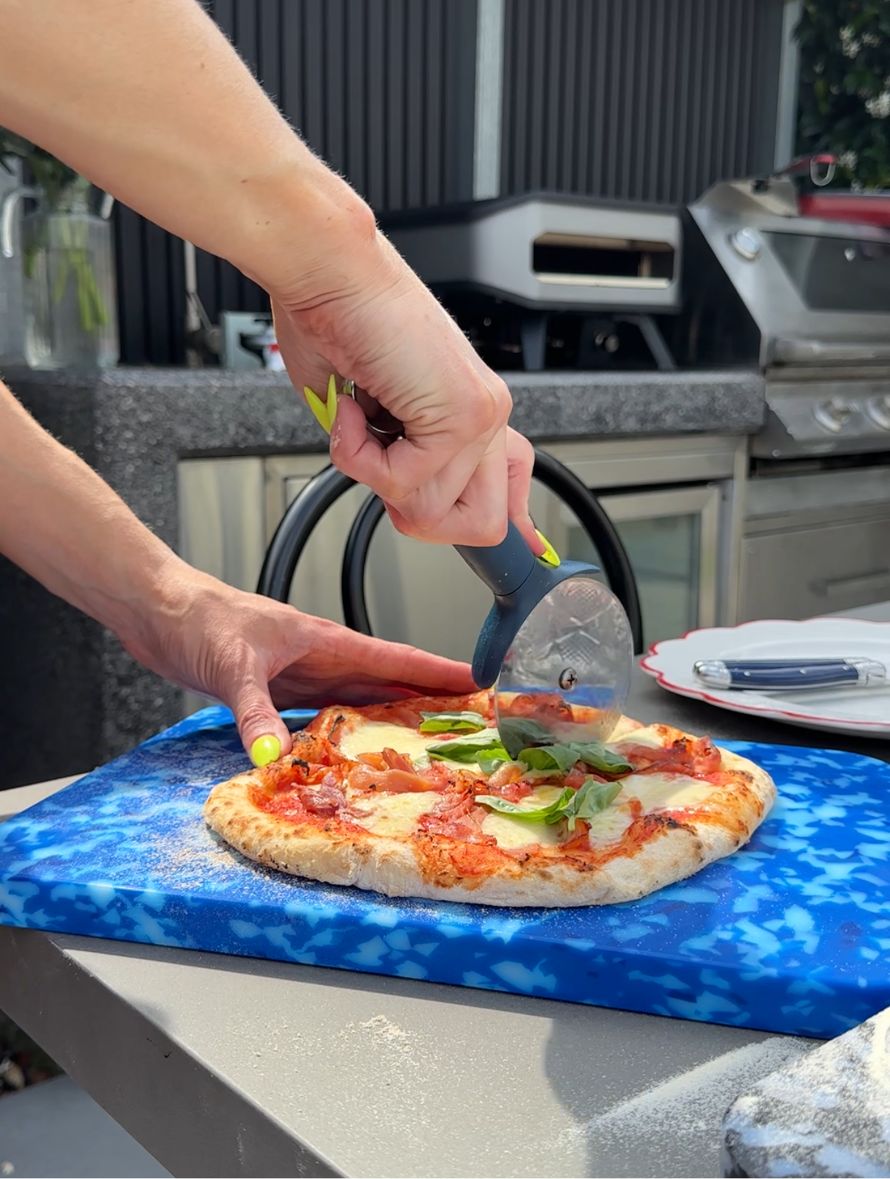 Blue and white Pinchy chopping board used as pizza base while slicing fresh pizza outdoors on barbecue bench