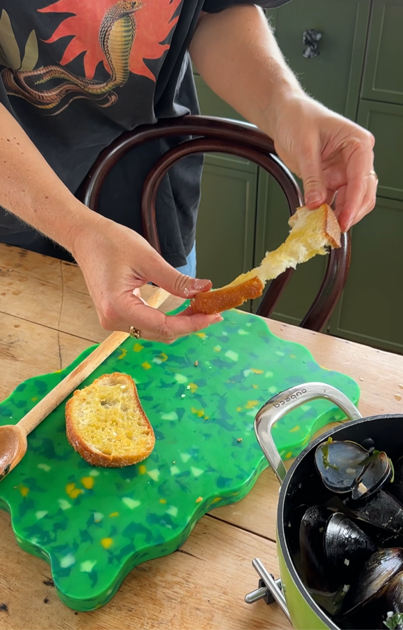 Green and yellow wavy Pinchy chopping board on wooden table with bread and pot of mussels – person tearing bread