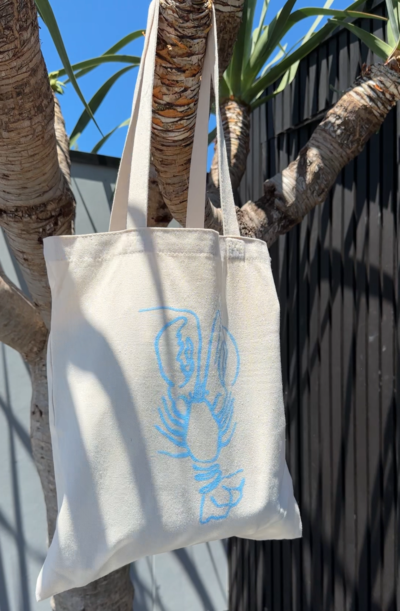 A beige cotton canvas tote bag with long straps, featuring a blue lobster print, hanging from a branch.