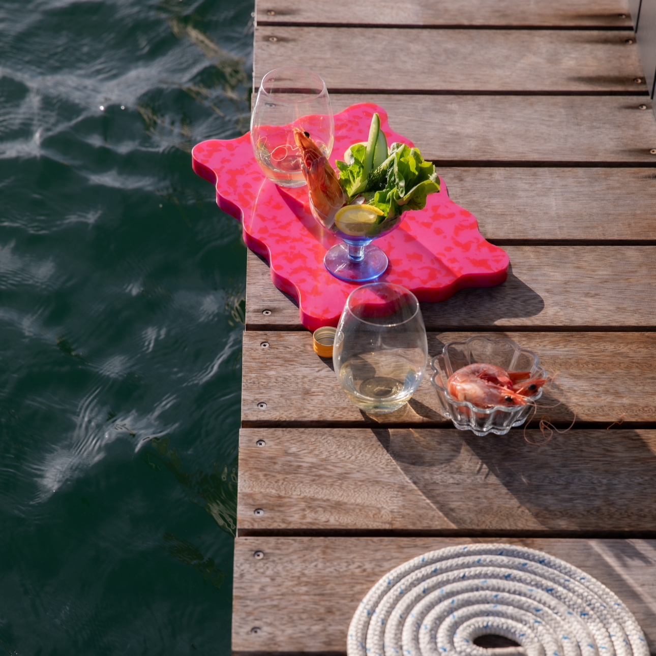 Pink and red wavy Pinchy board with prawns and wine on timber jetty beside the water