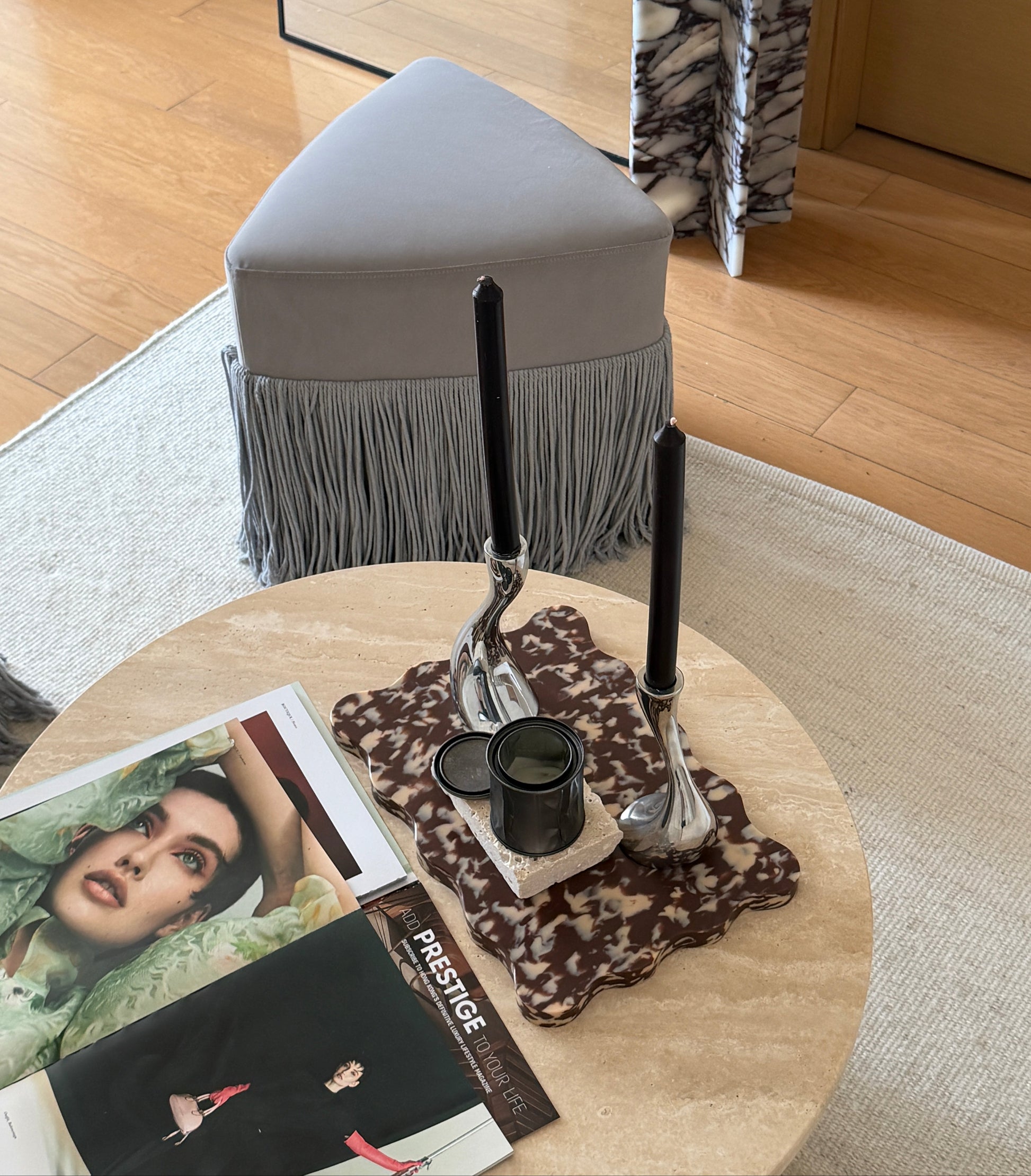 A styled interior scene showing a light gray upholstered ottoman with fringe, placed in a real room with wood flooring and a marble side table. On the foreground round table are magazines and a candle holder with black taper candles.
