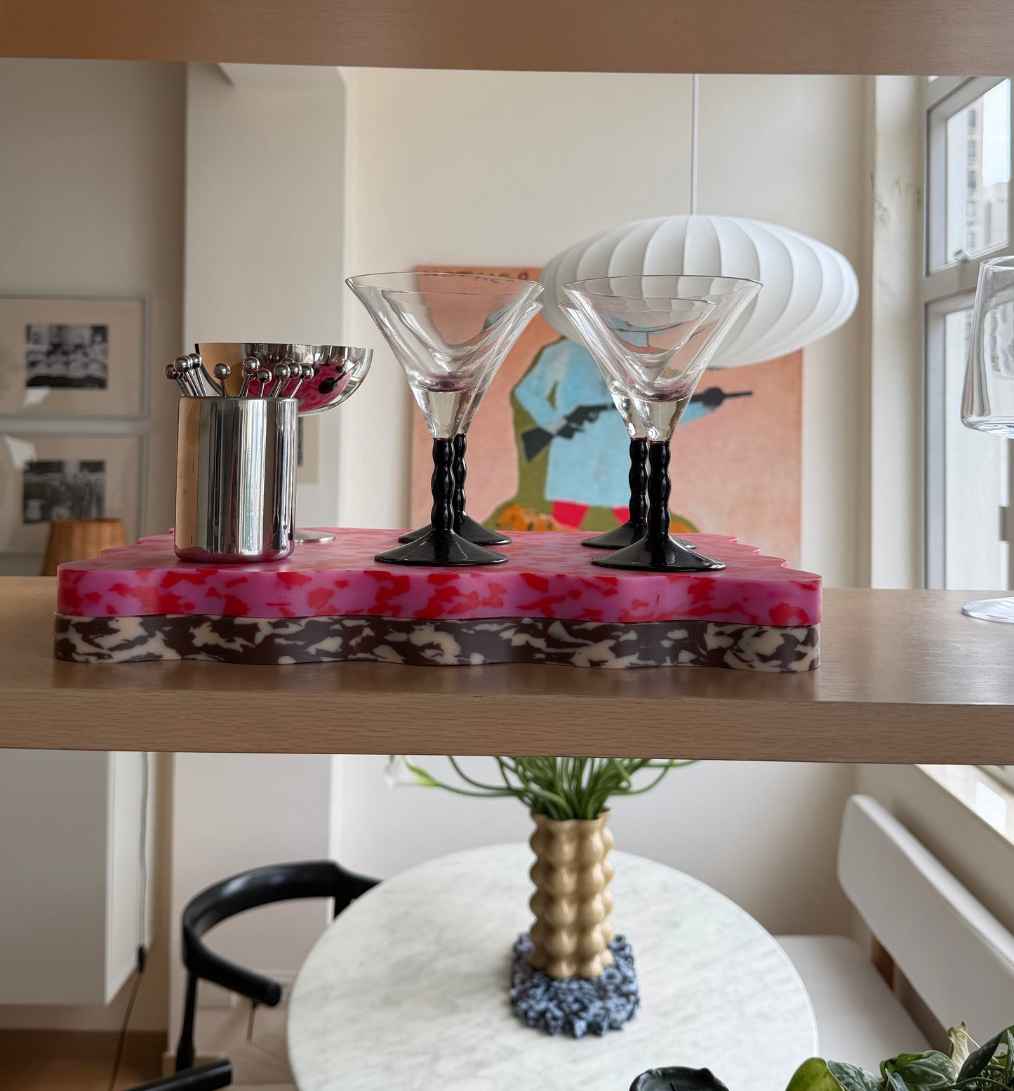 A decorative wavy cutting board with pink and red marbled top, holding two clear martini glasses with black stems and a small silver cocktail spoon holder, displayed on a wooden shelf in a bright interior.