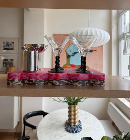 A decorative wavy cutting board with pink and red marbled top, holding two clear martini glasses with black stems and a small silver cocktail spoon holder, displayed on a wooden shelf in a bright interior.