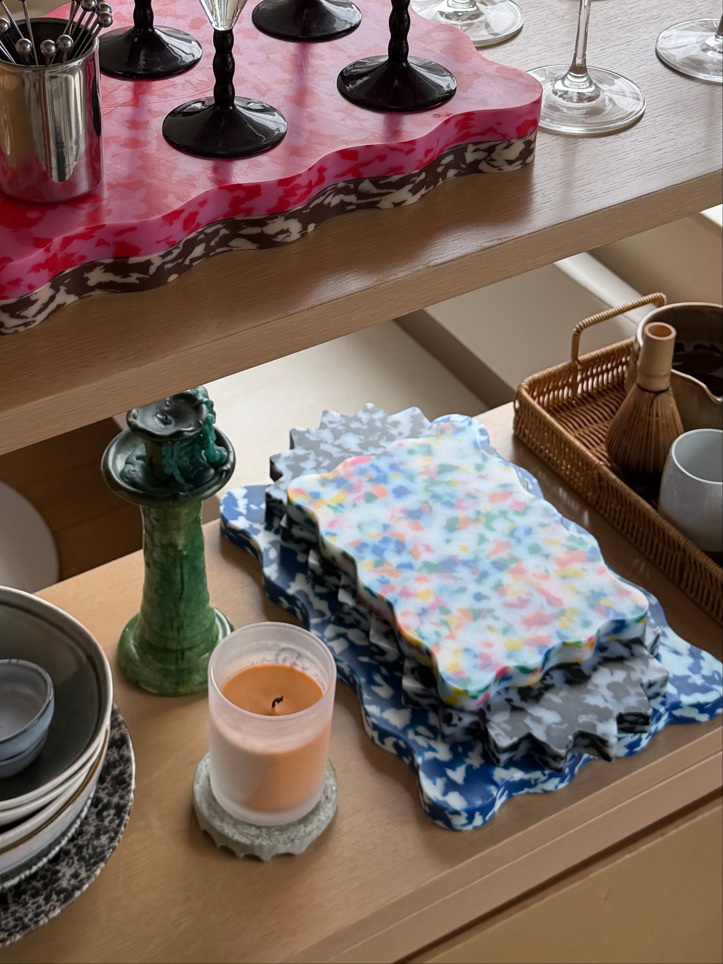 A styled interior shelf display featuring a pink marbled serving board with black martini glasses, clear wine glasses, stacked ceramic bowls, folded patterned napkins, and a candle in a real home environment.