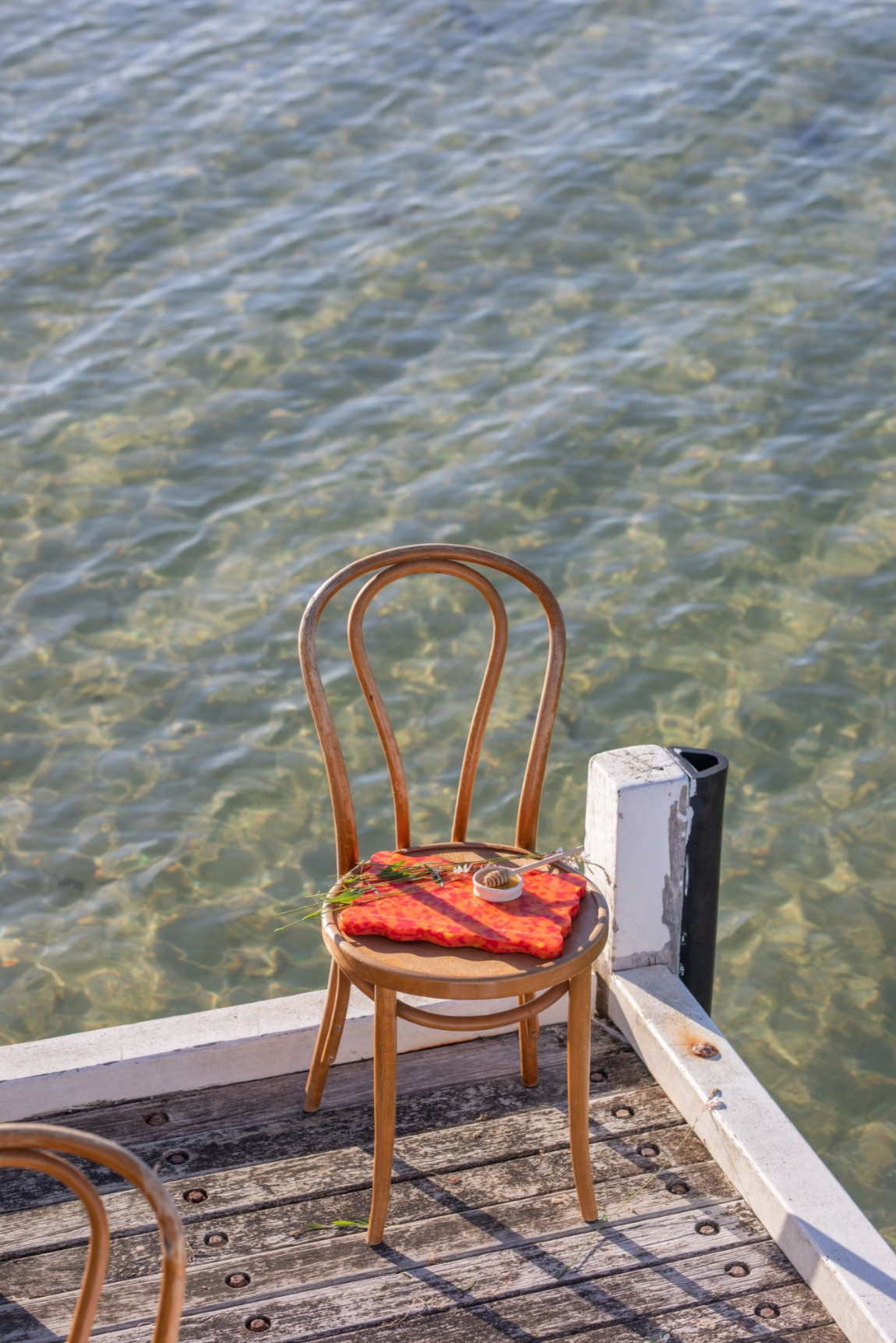 Orange and red wavy Pinchy chopping board on wooden chair by the water, styled with bread and herbs on jetty
