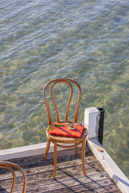Orange and red wavy Pinchy chopping board on wooden chair by the water, styled with bread and herbs on jetty