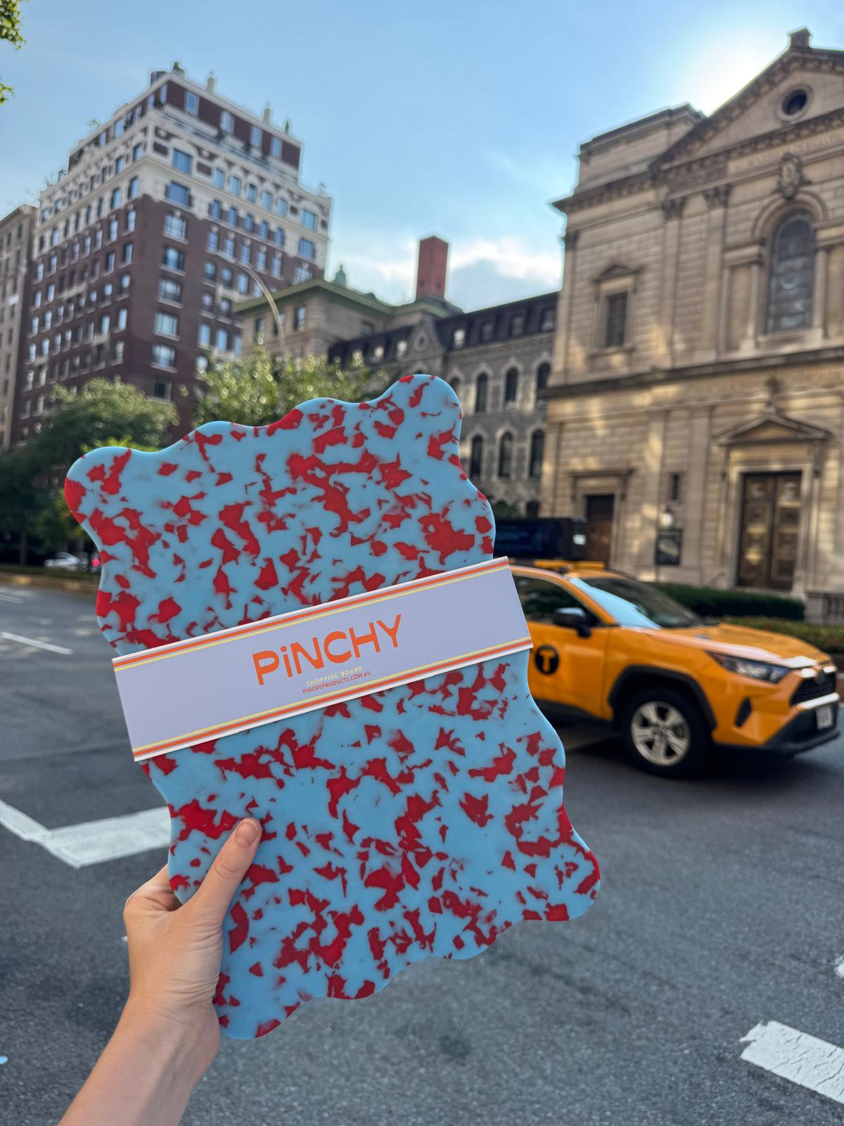 A hand holds a blue and red patterned, irregularly shaped board with a white band reading 'PINCHY' in orange text, with a city street, buildings, and a yellow taxi in the background.