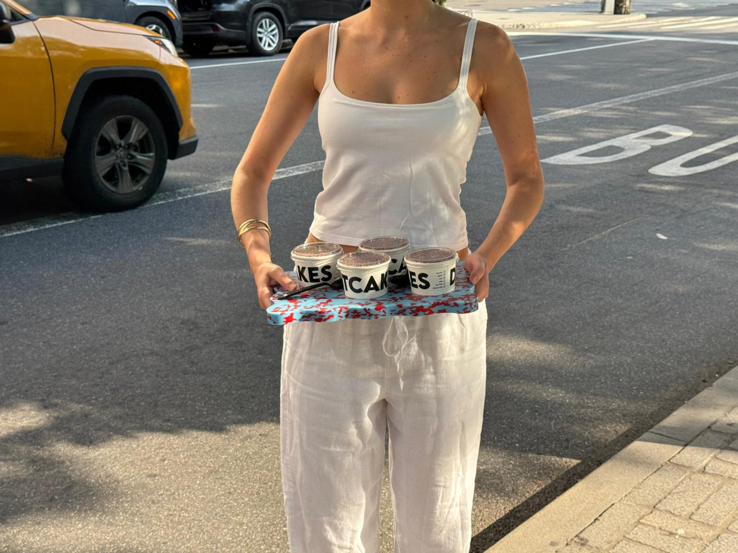 A woman in a white tank top and white pants stands on a NYC street holding a colourful wavy Pinchy tray with Dot Cakes on it. 